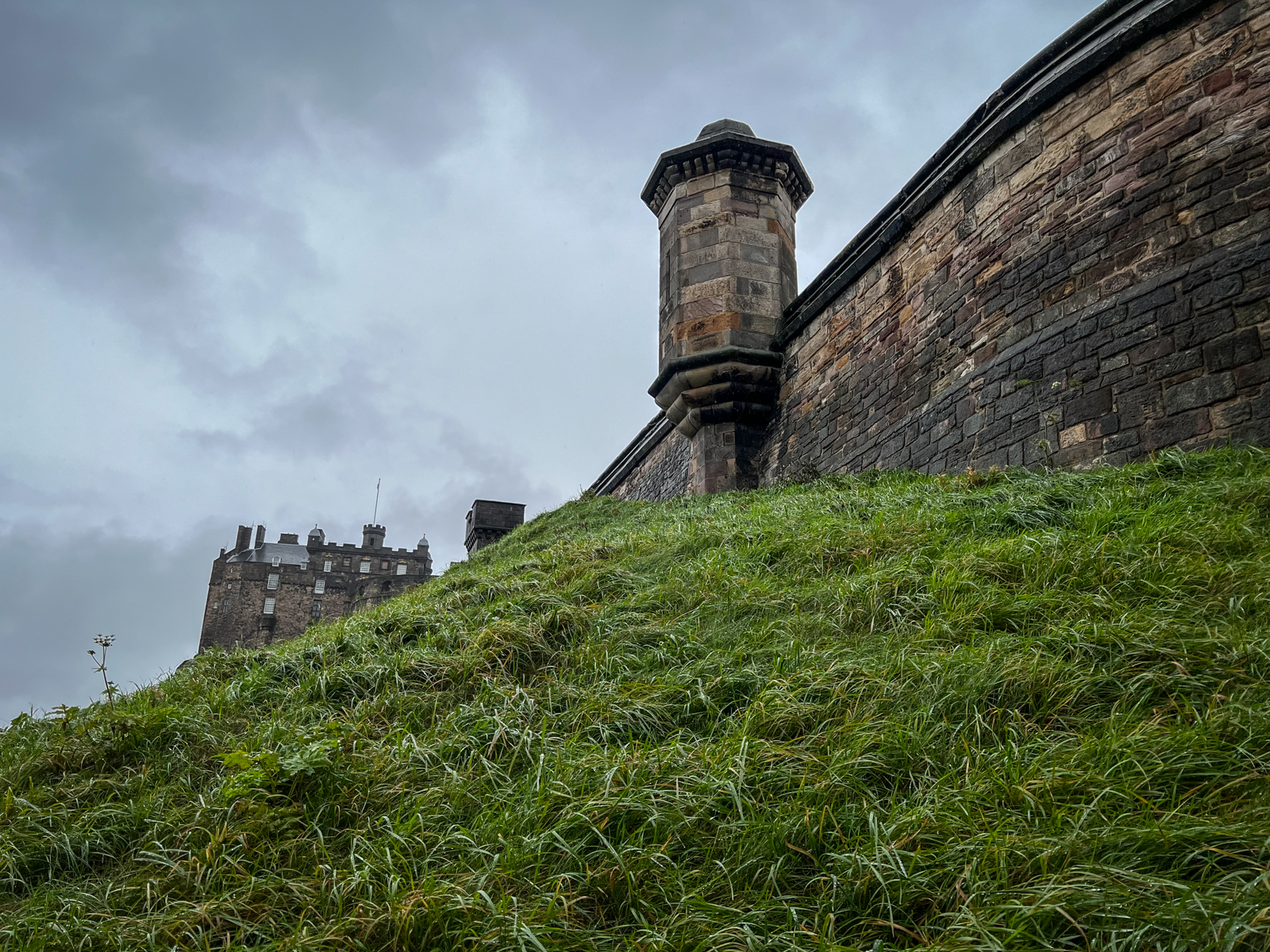 A grassy hillside leading up to a tall stone wall of a castle or fortification. In the background, a section of the castle peeks above the hill, set against a cloudy, overcast sky.