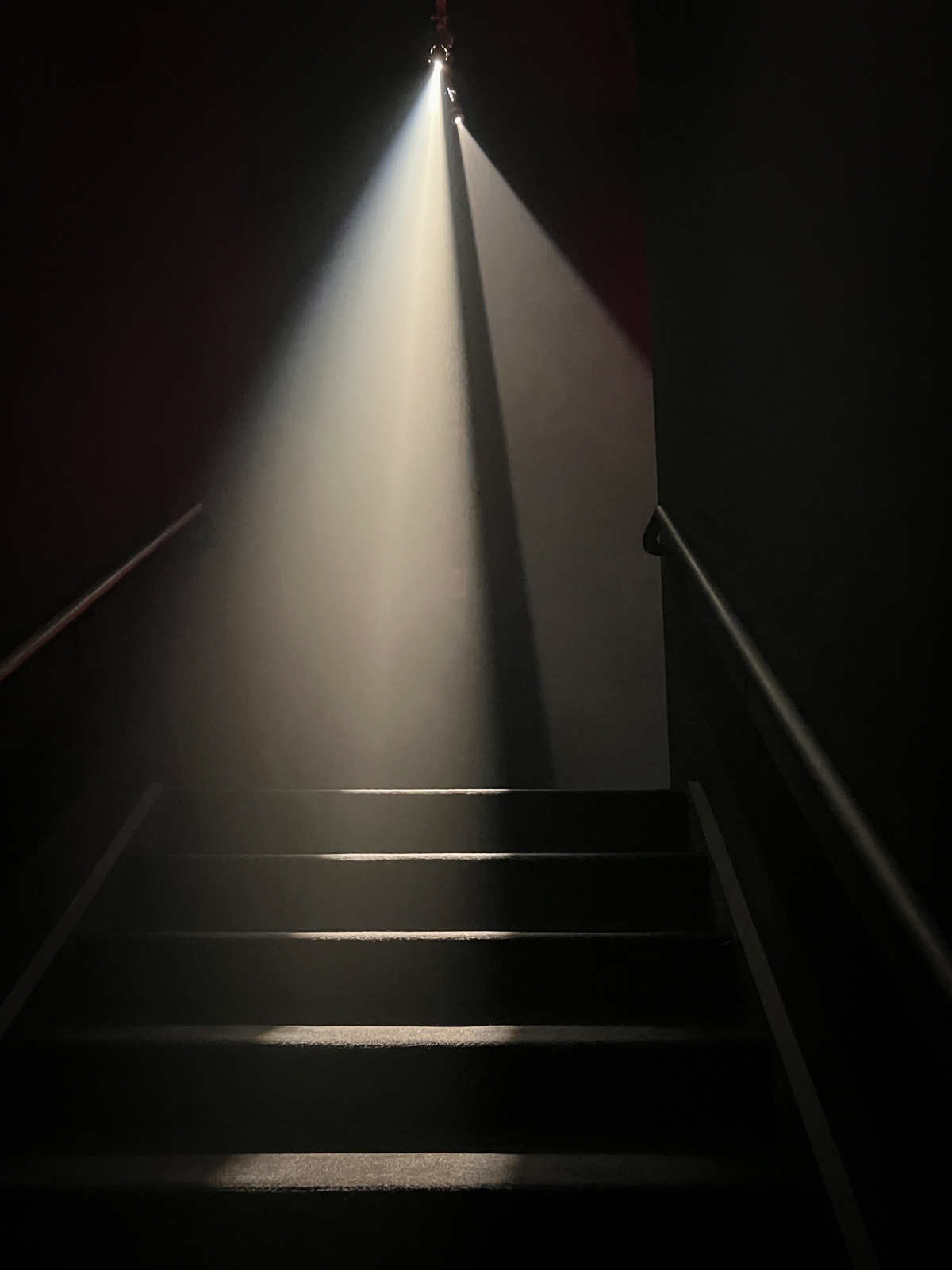A dark staircase illuminated by a single bright beam of light coming from above, casting sharp shadows and creating an ethereal, dramatic effect. The focus is on the contrasting light and shadow, emphasizing the texture and depth of the scene.