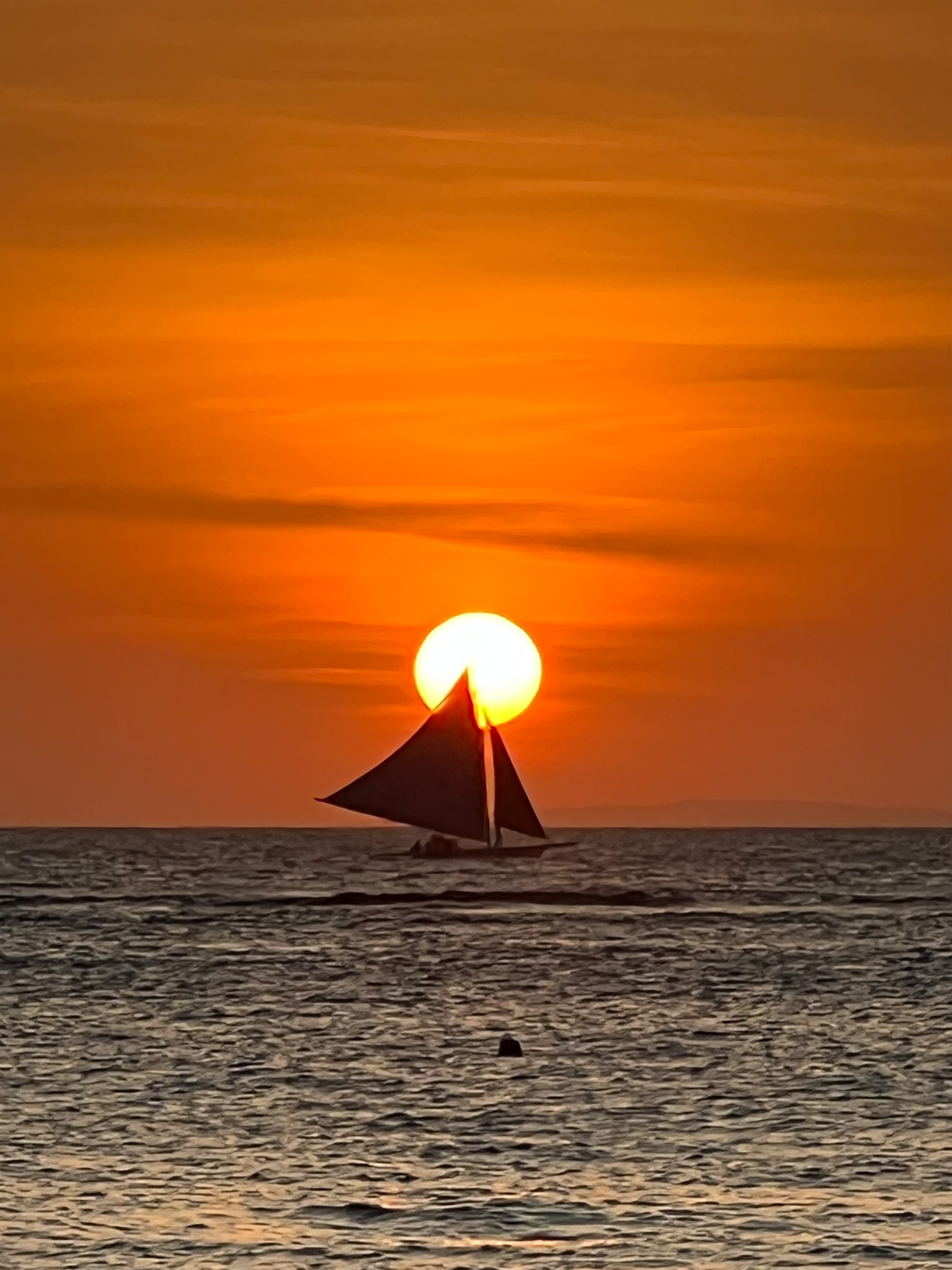 A sailboat silhouetted against a vibrant orange sunset, with the sun perfectly positioned behind its sail as it glides across calm ocean waters.