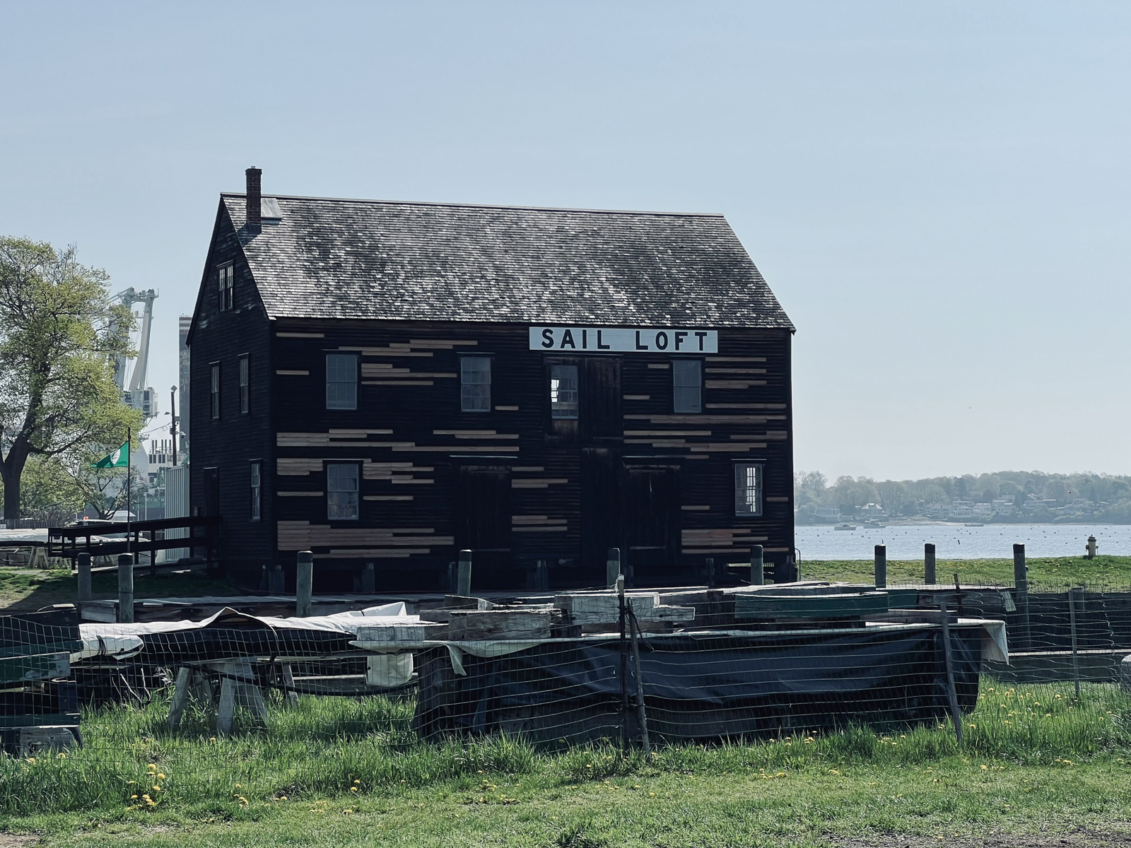 Historic wooden building labeled ‘Sail Loft’ near a waterfront, with scattered planks and materials in the foreground and a calm body of water behind it.