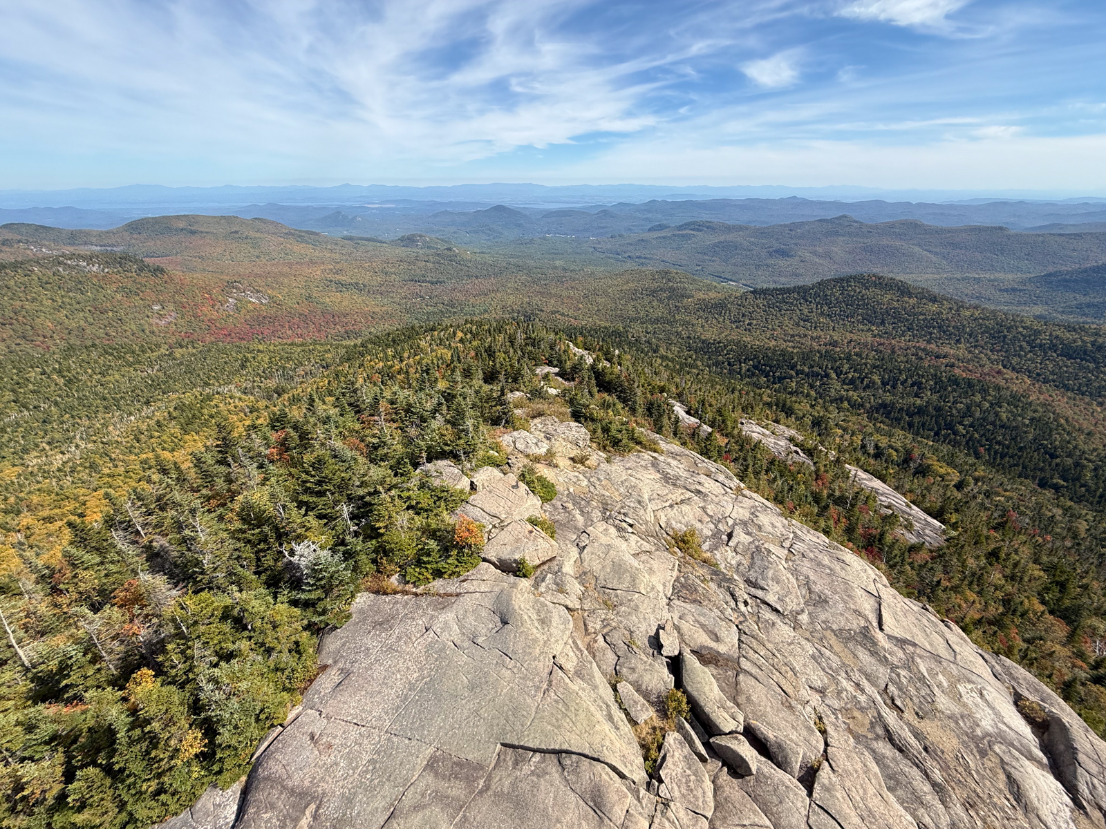 Expansive view from a rocky mountain summit, with rolling forested hills stretching into the distance under a blue sky streaked with wispy clouds.