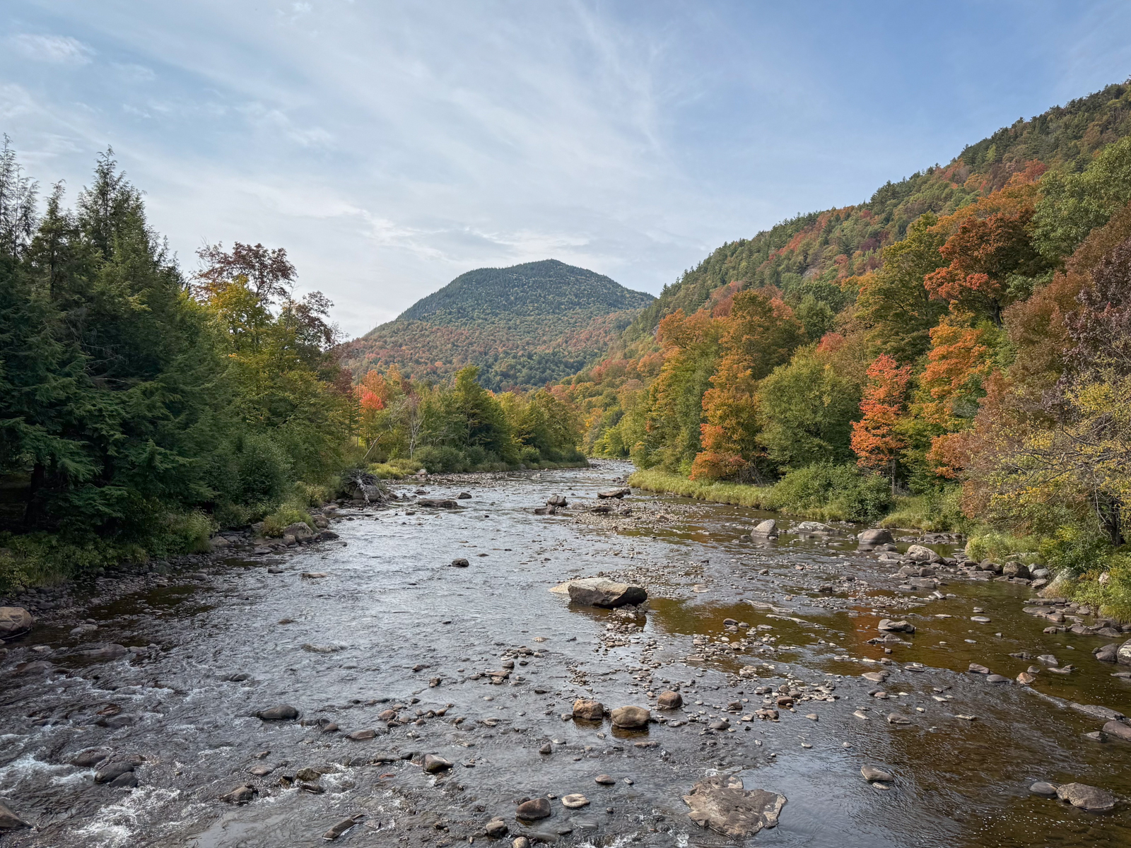 A rocky river winding through a valley of autumn-colored trees, with a green, forested mountain rising in the background.