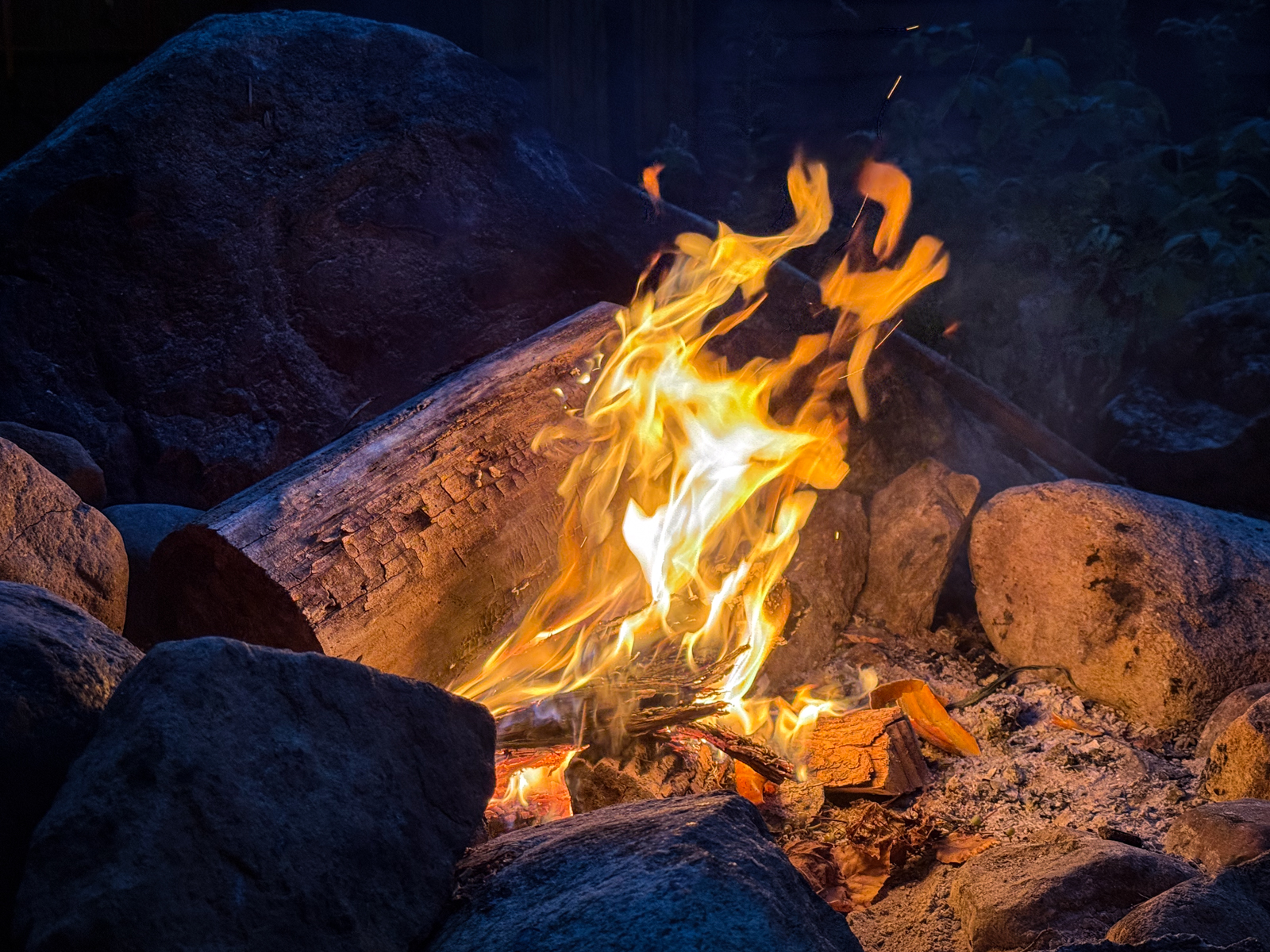 Bright orange flames rising from a log surrounded by large rocks in a nighttime fire pit.