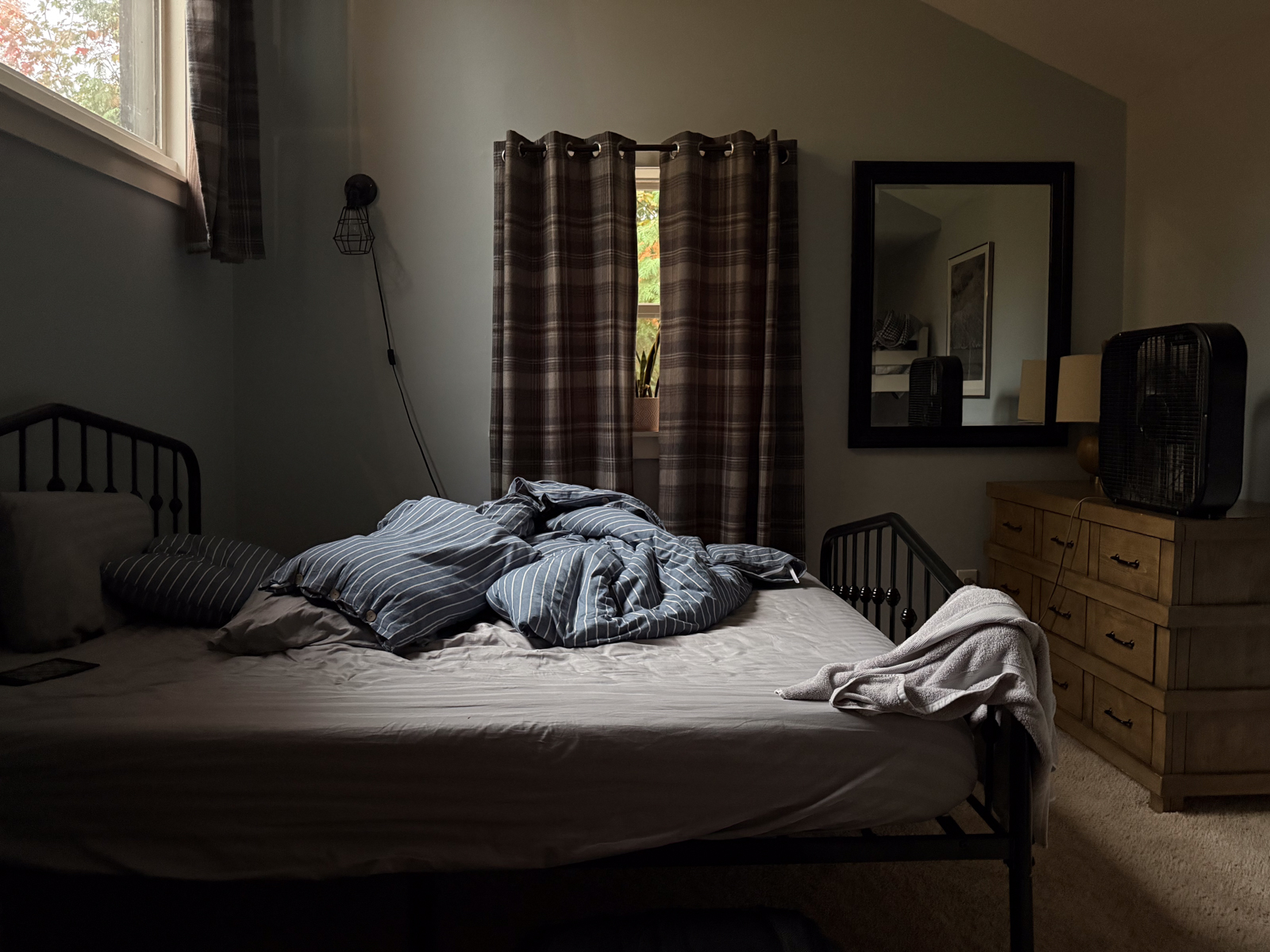 A dimly lit bedroom with an unmade bed of striped blue sheets, a dresser with a mirror, and curtains drawn across the window.