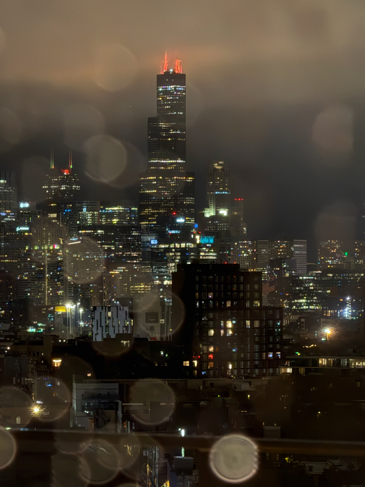 A nighttime cityscape featuring Chicago’s Willis Tower with its antennae glowing red against a cloudy sky. The skyline is brightly lit with numerous high-rise buildings, while large circular bokeh from raindrops or reflections blur parts of the foreground, creating a soft, atmospheric effect.